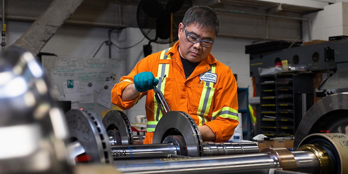 SkyTrain maintenance worker in orange safety gear working on train axle components in a workshop