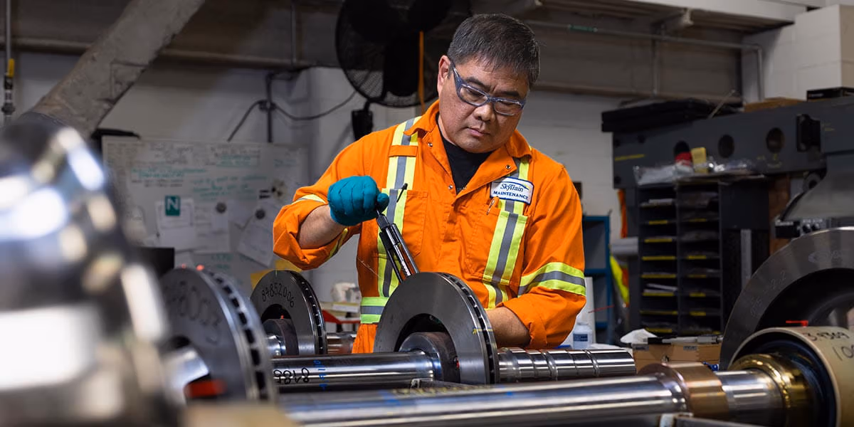 SkyTrain maintenance worker in orange safety gear working on train axle components in a workshop