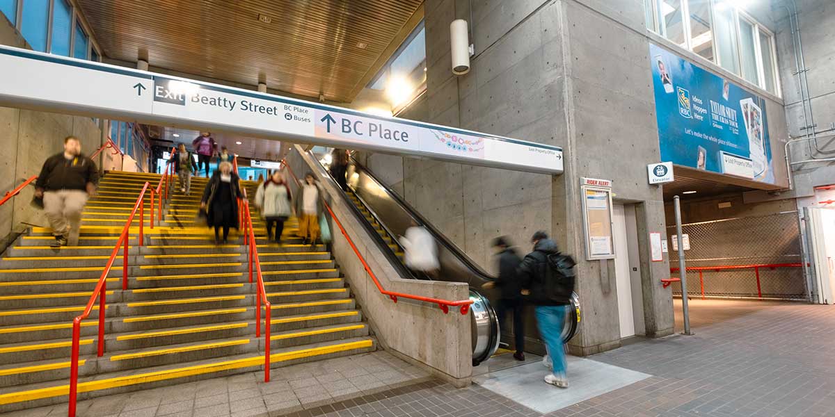 Busy Stadium-Chinatown SkyTrain station with people on stairs and escalator, signage pointing to Beatty Street and BC Place.