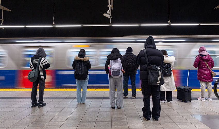 A group of bundled-up commuters waits on a brightly lit SkyTrain platform as a train speeds past.