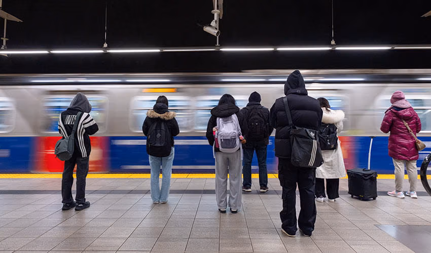 A group of bundled-up commuters waits on a brightly lit SkyTrain platform as a train speeds past.