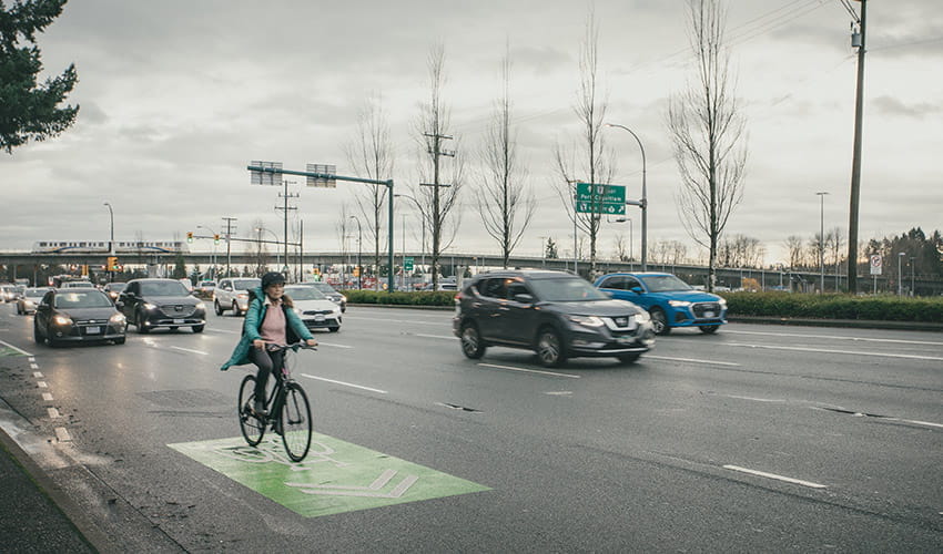 A women cyclist riding on a designated bike lane alongside moving traffic on a cloudy day, with cars and road signs visible in the background.