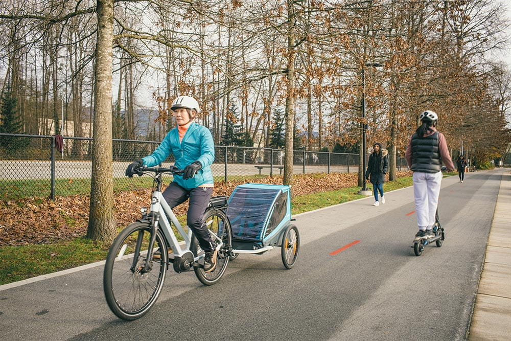 A woman riding an electric bike on a bike path while towing a children's bike trailer.